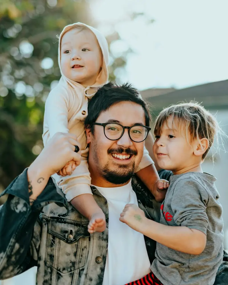 Pai sorrindo com os dois filhos, um deles sentado em seus ombros